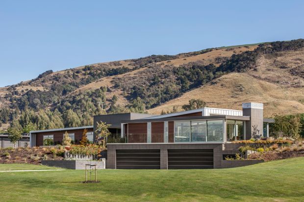 A modern single-storey home set against a dry, wooded hillside. The building features a low, angular flat roof, extensive floor-to-ceiling glazing, dark masonry or concrete walls, warm timber-toned cladding, and an integrated double garage. A broad manicured lawn and landscaped garden occupy the foreground.