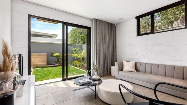Bright contemporary living room with a long grey upholstered sofa, rounded cream ottoman, black-framed glass coffee table, and floor-to-ceiling sliding doors opening to a landscaped courtyard. White painted brick walls, pale tiled flooring, full-height grey curtains, and dark-framed clerestory-style windows create a clean, refined interior.