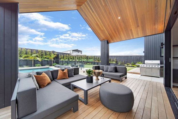 A sophisticated covered outdoor entertaining area with timber decking, a dramatic slatted timber ceiling and dark vertical-clad walls. The space features a large grey sectional sofa, coffee table and ottoman, with a built-in outdoor kitchen and barbecue at the rear. A swimming pool, landscaped garden and neighbouring hillside are visible beyond the seating area.