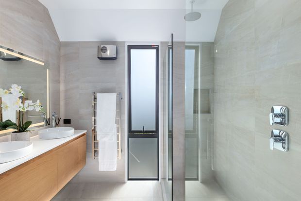 Bright contemporary bathroom with a double vanity and timber-look floating cabinetry, large-format pale stone-look wall tiles, frameless glass shower enclosure, wall-mounted chrome controls, heated towel rail, and a narrow frosted-glass window. A mirror reflects the vanity and orchid arrangement.