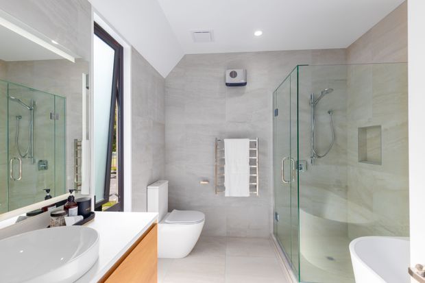 Bright contemporary bathroom featuring large-format light grey stone-look wall and floor tiles, a frameless glass walk-in shower, freestanding bathtub, wall-mounted toilet, timber vanity cabinetry, and a large mirrored wall.