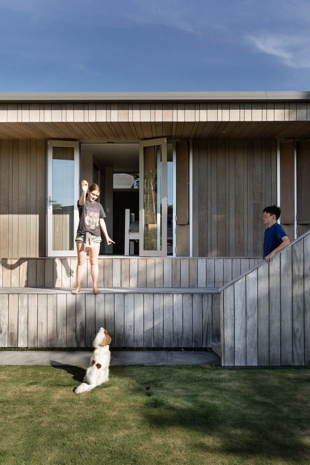 A low, contemporary timber-clad house viewed from the lawn, with a broad covered porch, elevated deck, open glazed entry, and a ramped or stepped access route with timber balustrades. Two people and a dog occupy the foreground, while the visible facade features vertical weatherboards, timber soffit lining, large glazed doors, and a shallow flat or low-pitched roofline.