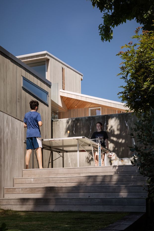 Exterior view of a contemporary multi-level timber-clad home with a broad stepped timber deck and concrete boundary wall. The building features clean rectilinear forms, projecting roof planes, large glazed openings, and warm timber soffit lining. People are playing table tennis on the deck, with mature trees framing the scene.
