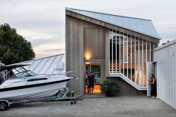 Exterior view of a contemporary timber-clad building with a steep asymmetrical metal roof and a tall glazed entrance facade featuring vertical timber fins and white framing. A boat is parked in the foreground beside the textured concrete driveway, with warm interior lighting visible through the entry.
