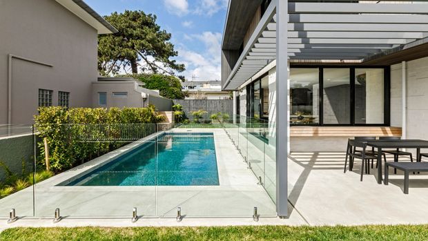 Contemporary residential courtyard with a narrow rectangular swimming pool bordered by frameless glass fencing. A covered outdoor dining terrace with a white pergola, timber soffit, large glazed openings, and pale masonry walls runs alongside the pool. The scene is bright and landscaped, with neighbouring residential structures visible beyond the courtyard.