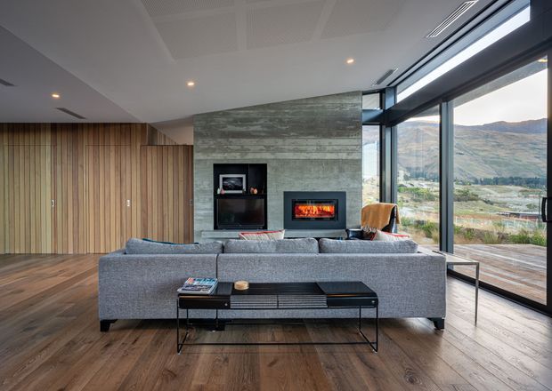 Contemporary living room with a large grey sectional sofa facing a built-in fireplace and television niche. The room features warm timber flooring, vertical timber wall panelling, a textured concrete fireplace wall, a slim black console table, recessed lighting, and expansive floor-to-ceiling glazing overlooking a mountainous landscape.