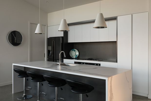 A contemporary kitchen dominated by a large island with a pale stone benchtop and integrated sink, accompanied by four black bar stools. White flat-panel cabinetry spans the rear wall, with a dark splashback and black appliances. Three pendant lights hang above the island, and a black circular wall clock adds a small accent. The room has a restrained monochromatic palette and clean-lined built-in joinery.