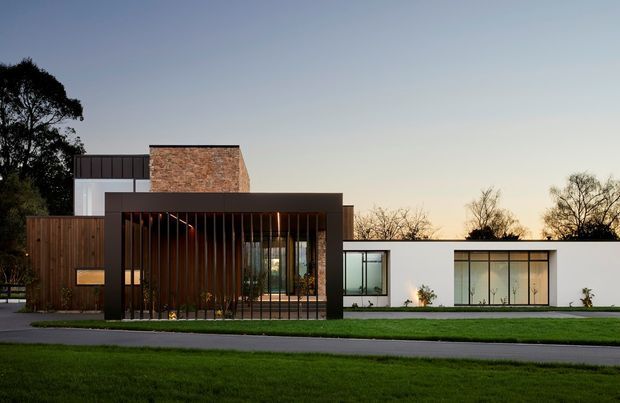 Contemporary single-storey residence viewed from across a suburban street, composed of crisp white rendered volumes, dark framed glazing, a projecting dark entry pavilion with vertical timber screening, and a prominent red-brown brick upper volume. A manicured lawn and mature trees surround the house.