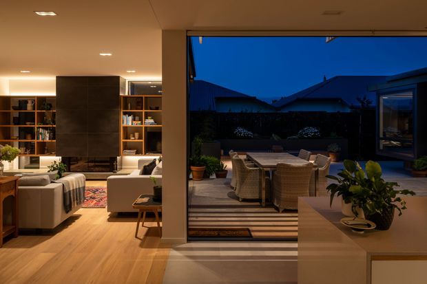 Warm evening interior looking from a contemporary living room through wide open glazing to a timber outdoor dining deck. The living area has light timber flooring, built-in dark joinery and shelving, upholstered seating, a low coffee table, and layered ambient lighting. The outdoor area contains a woven dining setting, potted plants, and landscaped garden beyond, with neighbouring rooflines visible in the twilight.