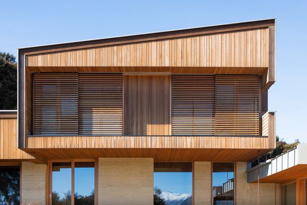 Exterior architectural photograph of a modern two-storey residence. The upper level features warm vertical timber cladding, deep overhangs, and broad windows screened by horizontal timber louvres. The lower level combines large glazed openings with light textured masonry walls, creating a strong timber-and-stone contemporary facade.