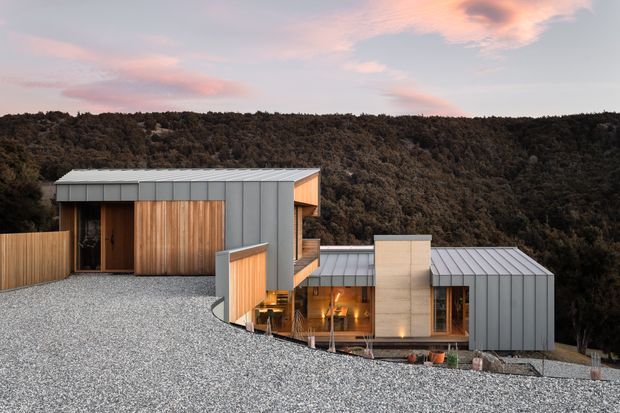 A contemporary multi-wing house integrated into a steep, wooded hillside. The building features low-pitched standing-seam metal roofs and grey metal cladding contrasted with warm vertical timber wall panels, glazed connections, and a prominent pale concrete central chimney or wall element. A broad gravel-covered foreground roof or terrace leads toward the forested landscape beyond, photographed at dusk.
