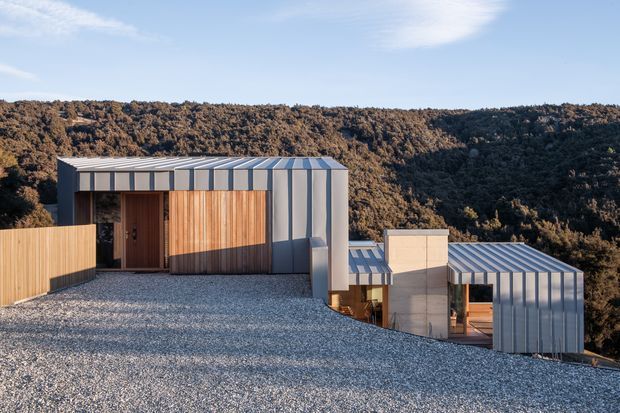 A contemporary hillside building composed of angular, low-profile volumes with corrugated metal roofing and cladding, warm vertical timber entry and facade panels, and concrete elements. The structure is set into a steep, wooded landscape, with a broad gravel-covered roof or terrace in the foreground and expansive bush-covered hills beyond.