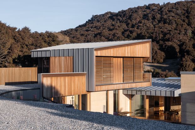 A contemporary multi-level house set into a wooded, hilly landscape. The building features a low-pitched metal roof, extensive vertical timber cladding, dark standing-seam metal wall panels, large glazed openings with timber louvred screens, and a gravel-covered foreground roof or terrace.