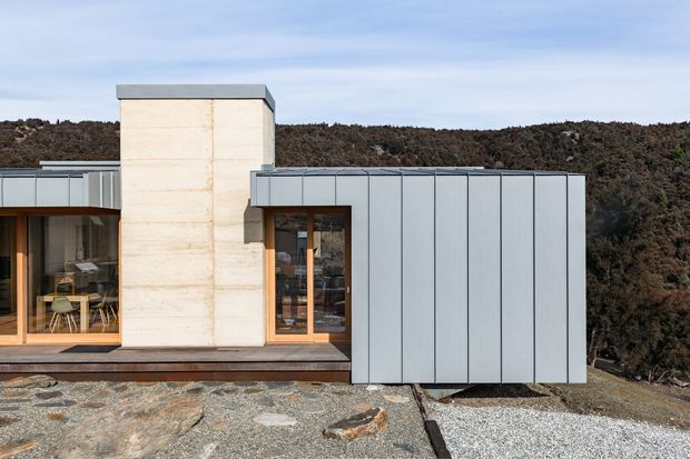Exterior view of a low, modern dwelling set against a rugged natural hillside. The facade combines pale board-formed concrete, large timber-framed glazed doors, dark timber decking, and a projecting volume clad in vertically panelled light-grey metal. The building has a flat roof and strong rectilinear massing, with exposed rock and gravel in the foreground.