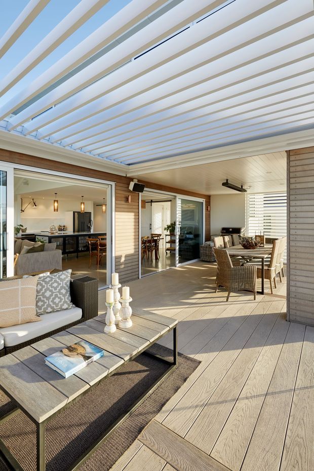 A bright covered outdoor terrace or patio with a large adjustable louvred pergola overhead, pale wood-look decking, timber-clad walls, and sliding glass doors opening into an indoor living and dining area. The terrace is furnished with woven lounge seating, a coffee table, and an outdoor dining set, creating a contemporary indoor-outdoor entertaining space.