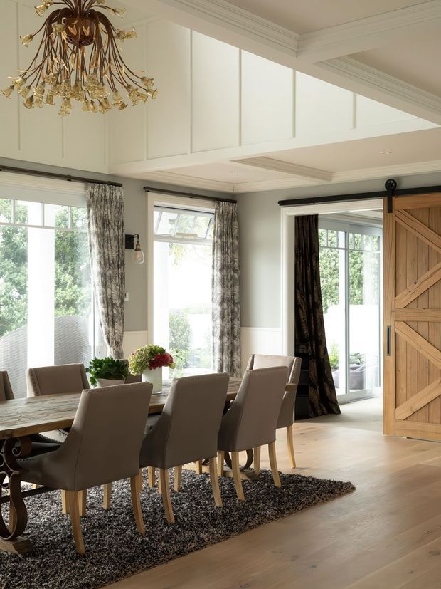 A bright, elegant dining room with a large timber dining table surrounded by upholstered dining chairs on a dark textured rug. The room features pale painted panelled walls, elaborate ceiling cornices and boxed detailing, tall windows with patterned curtains, light timber flooring, and a large rustic sliding barn door with diagonal bracing. An ornate gold chandelier provides a decorative focal point.