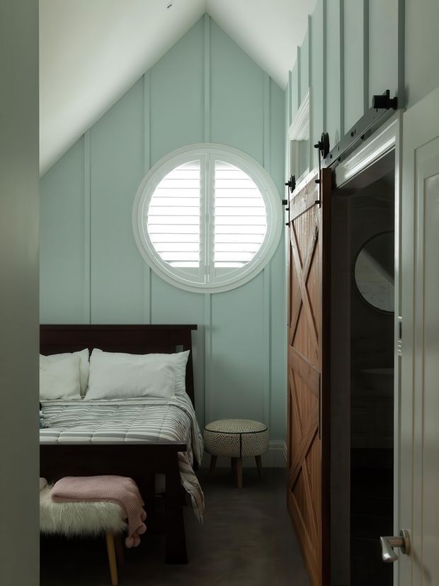 A compact, narrow bedroom with a steep vaulted ceiling, pale sage-green vertical wall panelling, and a round shuttered window. A dark timber bed with white bedding occupies the left side, while a small upholstered stool and bench add seating. On the right is a timber barn-style sliding door opening toward a dark adjoining space, with a white hinged door in the foreground. The room has a calm, tailored coastal feel with strong architectural detailing.