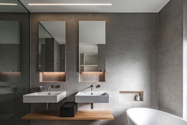 Contemporary bathroom with two wall-mounted white basins, mirrored medicine cabinets, a long timber vanity shelf, large-format dark grey wall tiles, and a freestanding white bathtub. Linear integrated ceiling lighting and recessed wall niches create a clean, refined fit-out.