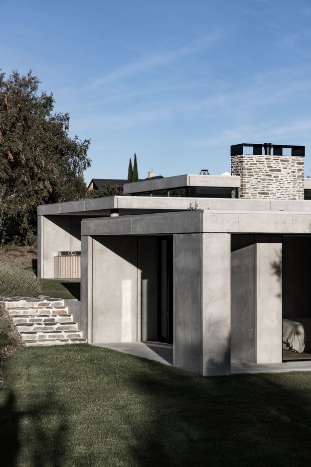 Exterior view of a low-slung contemporary concrete residence set into a landscaped site. The building features broad flat roof planes, deep projecting slab edges, dark recessed glazing and doors, exposed smooth grey concrete walls, and a prominent rough stone chimney or upper wall element. Stone steps and retaining walls connect the lawn to the entry, with mature planting framing the house.