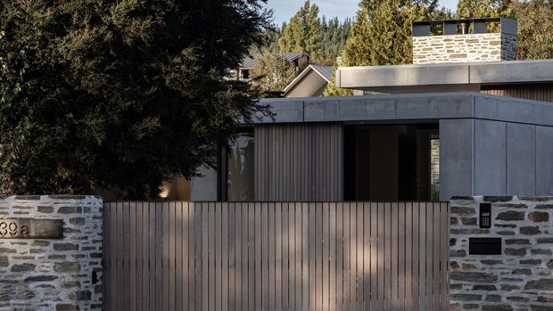 Street-facing view of a low-profile contemporary residence, partially screened by mature trees. The façade combines dark vertical timber cladding, large dark-framed glazing, smooth grey wall panels, and a low stone boundary wall with a matching stone chimney or rooftop feature. A tall timber privacy gate and fence run across the foreground, while the flat roofline and restrained material palette create a modern, private architectural expression.