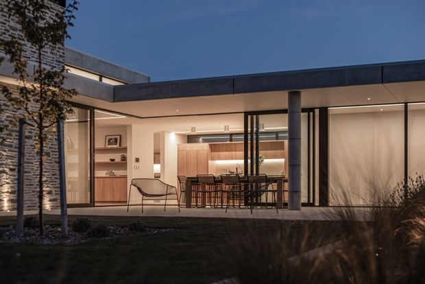 Twilight exterior view of a sophisticated single-level residence with a broad flat roof and an open, glazed entertaining area. The sheltered terrace reveals a warm contemporary kitchen and dining space with timber cabinetry, a dining island, bar stools, shelving, and minimalist furnishings. Dark metal-framed glazing, a concrete column, textured stone wall, and carefully planted landscaping create a refined indoor-outdoor connection.