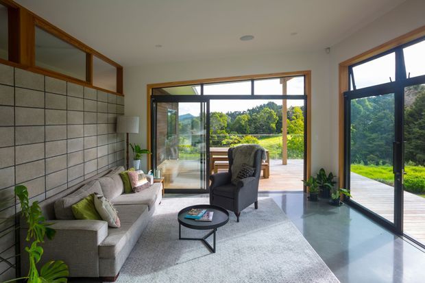 Bright contemporary living room with floor-to-ceiling black-framed glazing opening onto a timber deck and landscaped garden. The room features a large pale grey sectional sofa, dark armchair, round black coffee table, grey rug, indoor plants, and a distinctive tiled feature wall with timber-framed clerestory glazing.