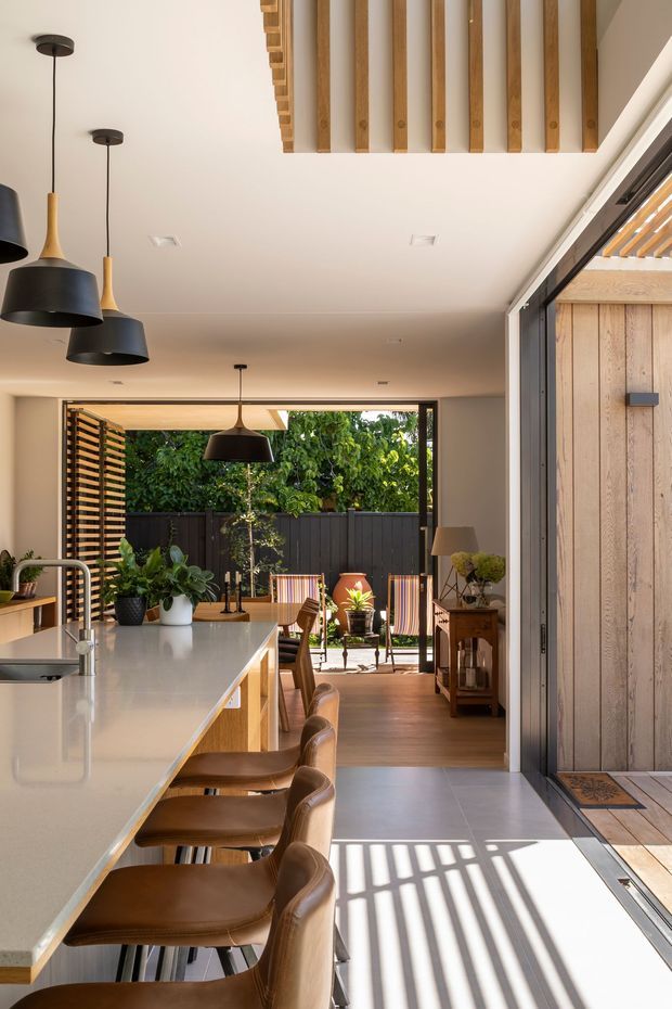A bright, open-plan contemporary kitchen and dining space viewed toward a leafy outdoor patio. The kitchen features a long light stone island with timber cabinetry and several brown upholstered stools, black pendant lights, and extensive glazed sliding doors. Warm timber flooring continues into the dining area, while a timber-lined entry or exterior wall and exposed timber ceiling battens add texture. Strong sunlight creates geometric shadows across the pale floor.