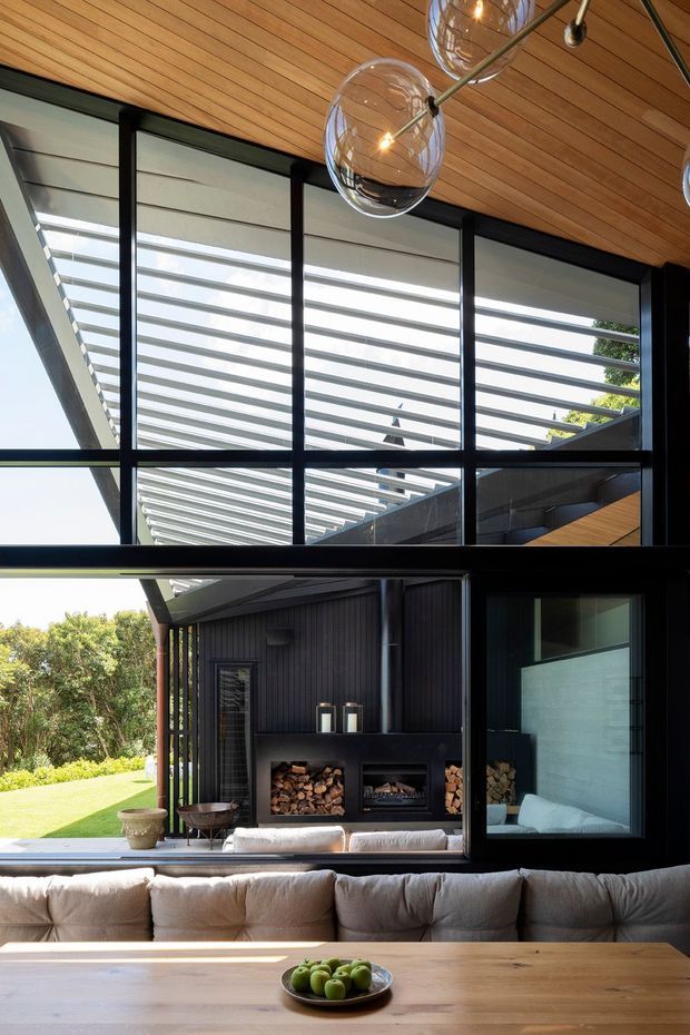A sophisticated open-plan living area with a timber-lined ceiling, expansive black-framed glazing, and a strong indoor-outdoor connection. The room features a large upholstered sofa, timber dining table, sculptural glass pendant lights, and a dark built-in fireplace wall with stacked firewood. A covered outdoor structure and landscaped garden are visible beyond the glazing.