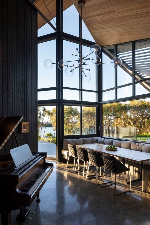 A dramatic double-height dining and living space with a grand piano, long dining table, upholstered bench seating and quilted bar stools. Floor-to-ceiling black-framed glazing wraps the room, opening views to a landscaped garden and water beyond. The ceiling is clad in warm timber boards, while a sculptural globe pendant hangs centrally above the table. The interior combines dark vertical wall panelling, polished concrete flooring, expansive glazing and strong indoor-outdoor connection.
