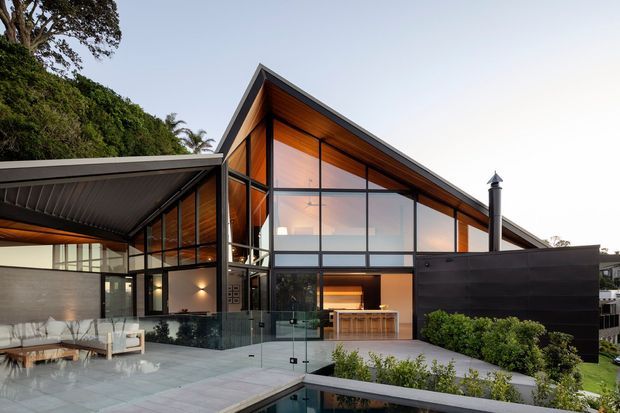 Exterior architectural view of a contemporary split-level residence with dramatic pitched roof forms, extensive floor-to-ceiling glazing, dark metal framing, warm timber-lined soffits, and an outdoor terrace overlooking a narrow pool. Dense greenery borders the home, while the glazed ground floor reveals a kitchen and living area inside.