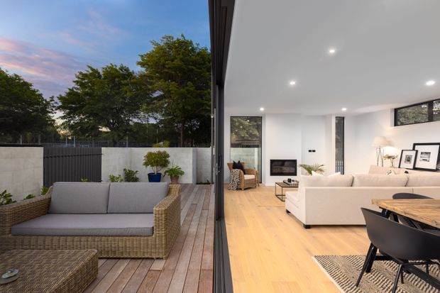 Contemporary open-plan living and dining interior opening onto a timber deck through extensive floor-to-ceiling glazing. The bright white room has pale timber flooring, a large cream sectional sofa, fireplace, framed artwork and a timber dining table with dark chairs. The outdoor deck includes woven rattan-style seating, raised planters and a dark privacy fence, with mature trees beyond.