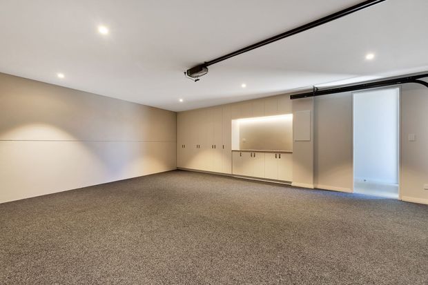 Spacious, clean-lined garage interior with a broad carpeted floor, integrated cream storage cabinetry and a softly illuminated recessed display niche along the rear wall. The white ceiling includes recessed downlights and a visible sectional garage-door track, while a frosted-glass pedestrian door opens on the right.