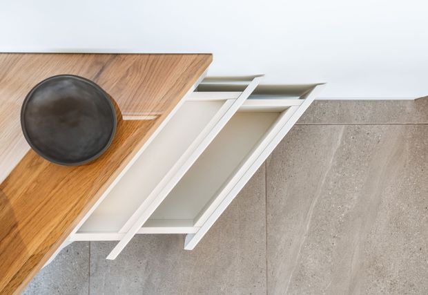 Top-down detail of a minimalist built-in storage unit with several white handleless drawers pulled open beneath a warm timber countertop. A dark round bowl sits on the benchtop, and large light-grey stone-look floor tiles surround the joinery.
