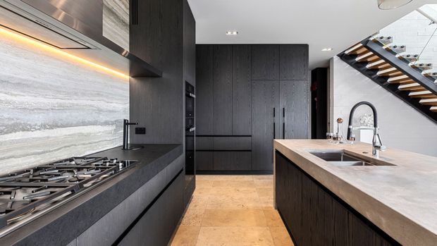 A sophisticated modern kitchen with dark vertical-grain cabinetry, a large stone island with an integrated sink, a gas cooktop, and a full-height stone splashback. Warm timber flooring and an open staircase with timber treads and a glass balustrade add contrast to the predominantly charcoal and grey palette.