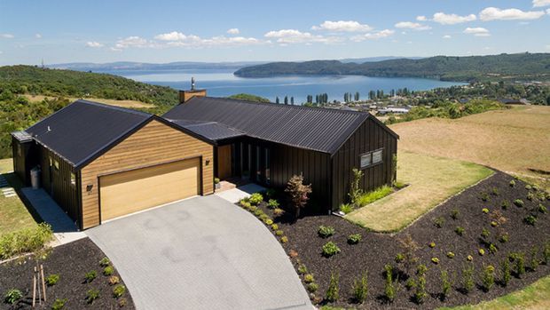 Aerial view of a contemporary single-storey hillside home with dark standing-seam metal roofing and dark vertical cladding, complemented by a warm timber-clad garage wing. The property has a broad concrete driveway, landscaped planting beds, and expansive views across a lake and surrounding rural hills.