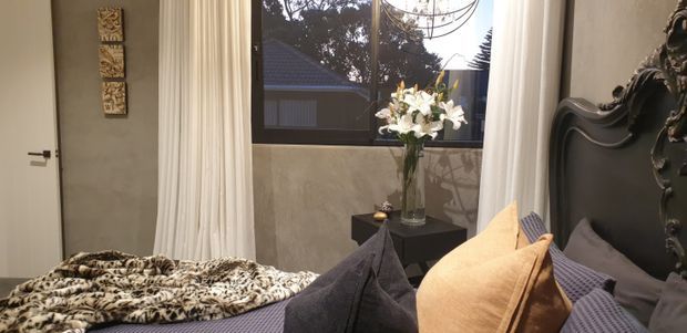A moody, elegantly styled bedroom with a large upholstered bed, layered dark and mustard cushions, a patterned throw, floor-to-ceiling curtains, a black bedside table with flowers, and an ornate dark carved feature element at the right. A large window overlooks neighbouring rooftops and trees.