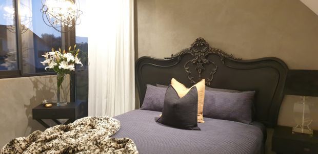 A richly styled bedroom with a dark ornate carved bedhead, coordinated mauve-grey bedding and cushions, sheer white curtains, a sculptural chandelier, bedside tables, and a floral arrangement. The room has a dramatic, elegant ambience with textured neutral walls and a view through the window.
