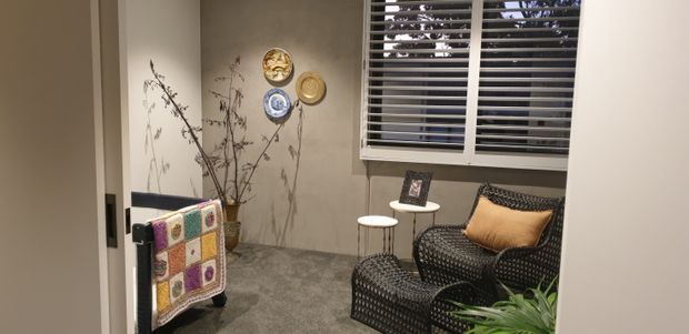A compact, warmly styled sitting nook or reading area with a dark woven armchair and matching ottoman, small round side tables, a textured grey carpet, neutral taupe walls, plantation shutters, decorative wall plates, and a colourful textile draped over a nearby rail. A leafy plant is visible in the foreground.