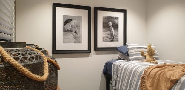 A cozy compact bedroom with a single bed dressed in blue, grey, and white striped bedding, a tan throw, and decorative pillows. Framed black-and-white family photographs hang on the light neutral wall, while a woven basket with rope handles sits in the foreground beside white plantation shutters.