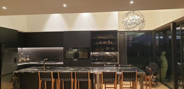 A sophisticated open-plan kitchen and dining area with matte black cabinetry, dark stone island and benchtops, integrated appliances, glass-front display shelving, and a large dining island with timber-framed stools. Warm recessed lighting and a sculptural spherical pendant create a refined, dramatic atmosphere, while full-height glazing is visible on the right.