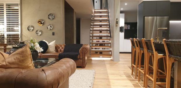 Open-plan living and kitchen interior with a warm, contemporary industrial character. A large cognac leather sectional sofa and matching leather armchairs occupy the living area, with a light textured rug and decorative wall plates. Timber flooring extends toward a central open-riser staircase with timber treads and dark metal supports. The kitchen features dark matte cabinetry, a stainless-steel refrigerator, and a long island or breakfast bar with multiple timber bar stools. Abundant daylight enters through timber Venetian blinds and a large window.