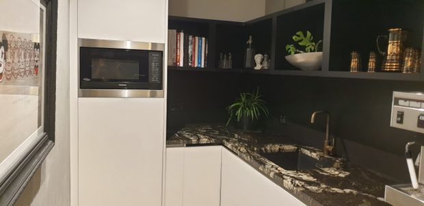 Compact contemporary kitchen with white flat-panel cabinetry, a built-in stainless-steel microwave, dark open shelving, and a dramatic dark patterned stone benchtop and splashback. A corner sink with a brass-toned tap sits beneath the shelving, with books and decorative items displayed above.