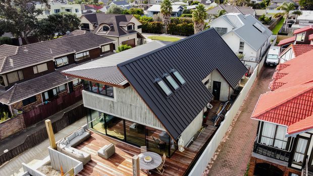 Aerial view of a contemporary multi-level residential home in a dense suburban neighbourhood. The house features a steep dark standing-seam metal gable roof with skylights, white and timber-toned exterior elements, extensive glazing, and a timber deck with outdoor seating. Adjacent homes, paved access paths, and established landscaping are visible.
