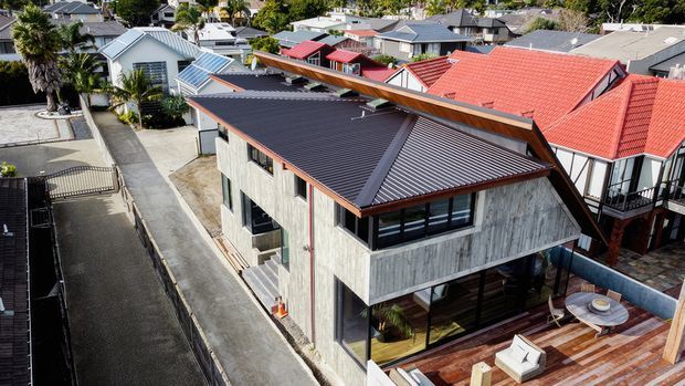 Aerial view of a contemporary multi-level residential house in a dense suburban neighbourhood. The home features a dark corrugated pitched roof with a prominent angled roof form, weathered vertical timber cladding, extensive dark-framed glazing, and a timber rooftop terrace or deck. Surrounding homes have varied pitched roofs and colourful metal roofing.