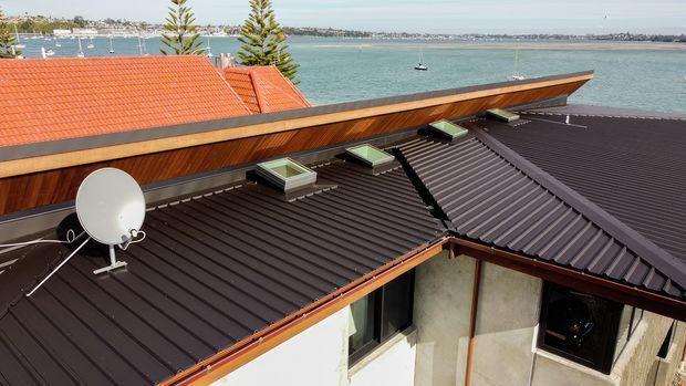Elevated exterior view of a low-pitched, multi-section metal roof with dark ribbed roofing, skylights, and a satellite dish. A timber-clad parapet or upper wall runs along the roof edge, with a terracotta-tiled roof and coastal harbour setting visible beyond.