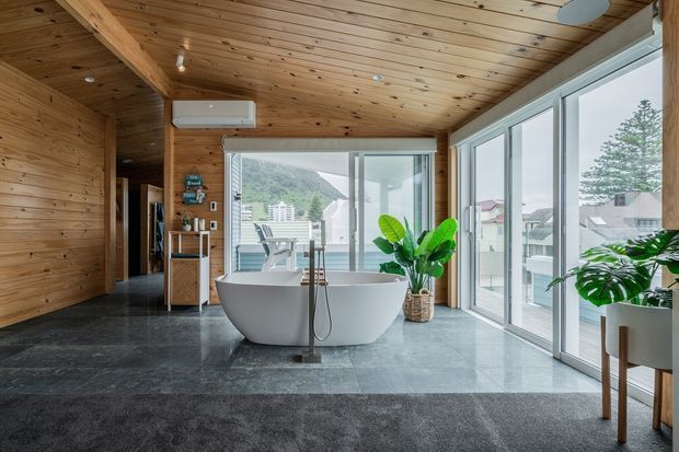 A contemporary open-plan bathroom with a freestanding white bathtub, dark stone-look flooring, extensive floor-to-ceiling glazing, and warm timber wall and ceiling lining. Indoor tropical plants and a timber vanity add natural texture, while the windows overlook a built-up hillside setting.