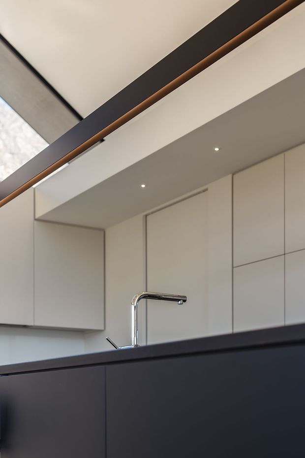 A close-up architectural view of a sleek contemporary kitchen, focused on the dark island cabinetry and chrome gooseneck tap. The kitchen features handleless white built-in cabinetry, a dark benchtop and island, recessed downlights, and a dramatic overhead linear architectural element with a warm timber or bronze-toned detail. The composition is minimalist and refined, with clean geometric lines and a neutral palette.