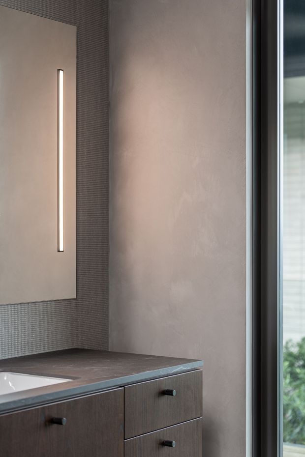 Close-up of a refined contemporary bathroom vanity featuring dark wood cabinetry with black pulls, a dark stone benchtop, a partially visible integrated basin, and a large frameless mirror with a slim vertical illuminated strip. The walls combine a small dark mosaic tile surface with a softly textured warm grey plaster finish, and a tall glazed opening brings in natural light.