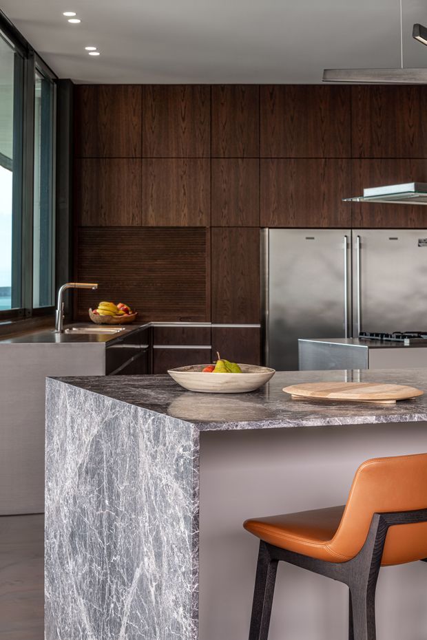 A sophisticated modern kitchen featuring dark timber cabinetry, stainless-steel appliances, a large grey veined stone island with seating, and a contrasting orange leather bar chair. Floor-to-ceiling glazing and recessed ceiling lighting bring natural light into the space.