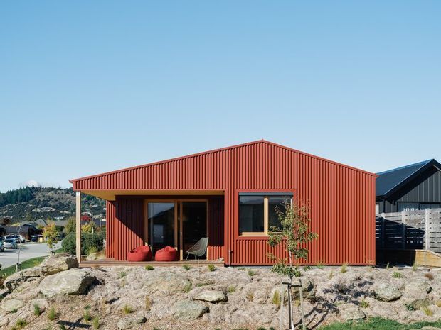 Single-storey contemporary house with a compact gabled form and deep red corrugated metal cladding. Large glazed openings sit beneath a covered entry porch, with a landscaped rocky foreground and neighbouring homes visible in the background.